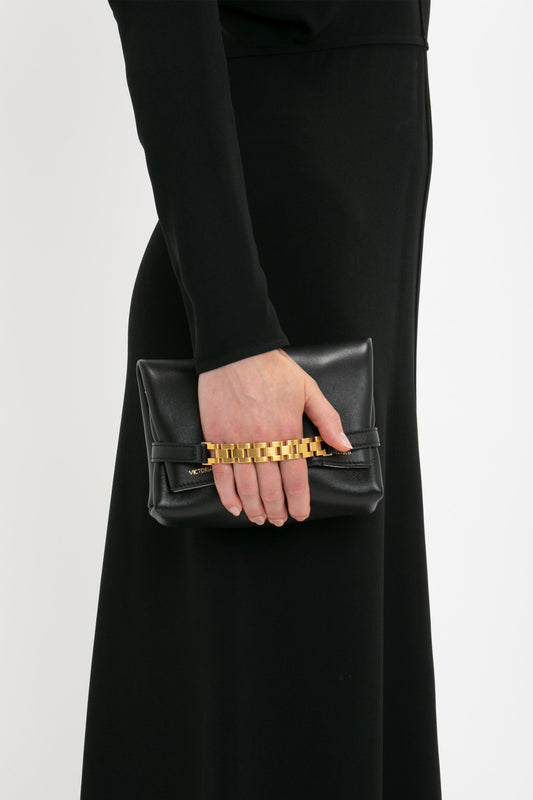 A person in a black Dolman Midi Dress by Victoria Beckham holding a small black clutch purse with a gold chain detail.