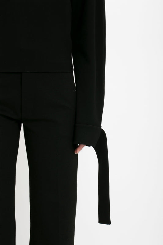 Close-up of a person wearing a Victoria Beckham Tie Sleeve Ponti Top In Black with extended cuff detail and matching black pants, standing against a white background.