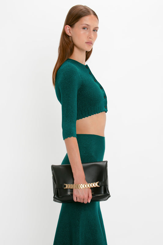 A woman with long hair, wearing a green crop top and skirt featuring pointelle stitch detailing, holds a large black clutch bag with gold accents in front of a plain white background. She is wearing the VB Body Cropped Cardi In Lurex Green by Victoria Beckham.