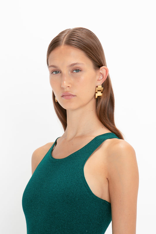 A woman with long, brown hair is wearing a VB Body Sleeveless Dress In Lurex Green by Victoria Beckham and large, geometric gold earrings, posing against a plain white background, showcasing a flared silhouette perfect for your new-season wardrobe.