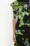 Close-up of a person wearing a Twist Shoulder Dress In Black Frost by Victoria Beckham with a green, black, and blue abstract pattern. The dress features ruffled shoulder detail and a stylish asymmetric hemline. Only the torso and part of the left arm are visible.