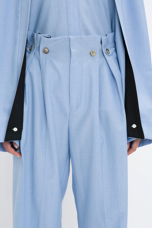 Close-up of a person wearing Victoria Beckham Gathered Waist Utility Trouser In Oxford Blue with button details. The person is also wearing a matching light blue jacket with black inner lining visible at the cuffs, epitomizing modern utility wear in Oxford Blue.