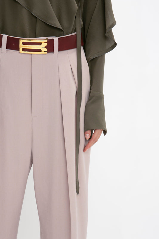 A person wearing Victoria Beckham's Double Pleat Trouser In Rose Quartz, a dark brown belt with a gold buckle, and a dark green blouse with a tie around the waist exudes a subtle masculine vibe.
