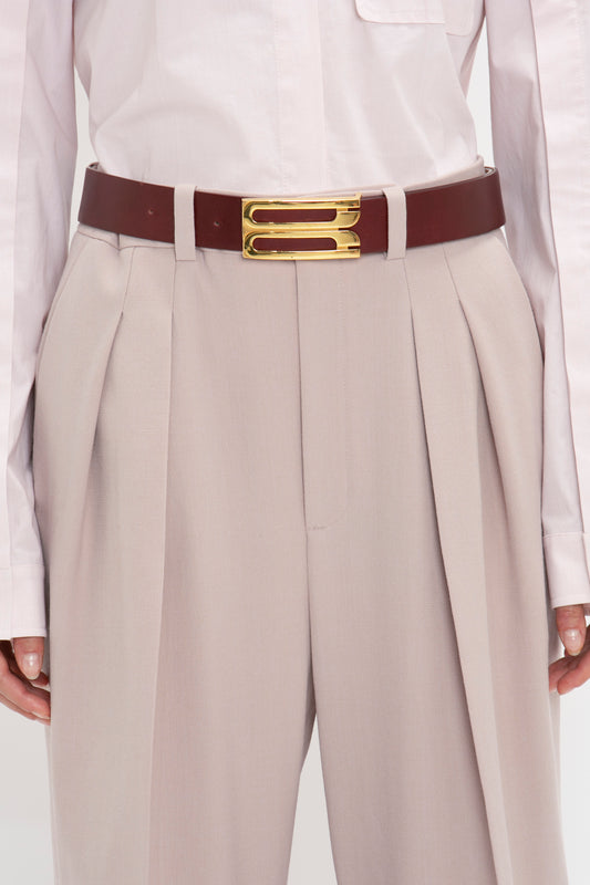 Close-up of a person wearing high-waisted beige trousers with pleats and a tucked-in white shirt, accessorized with a dark brown Victoria Beckham Jumbo Frame Belt In Burgundy Leather featuring gold hardware.