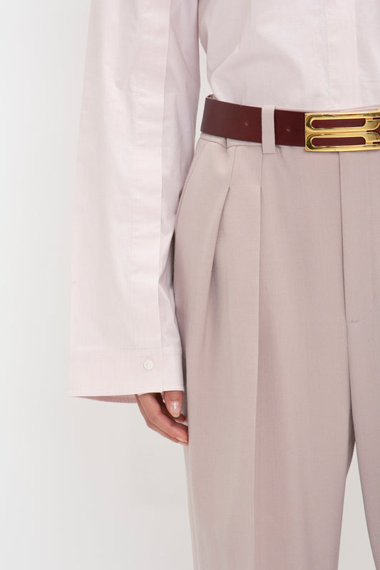 Close-up of a person wearing Victoria Beckham’s Button Detail Cropped Shirt In Rose Quartz tucked into beige high-waisted pants with pleats. The outfit is accessorized with a brown belt featuring a gold buckle.