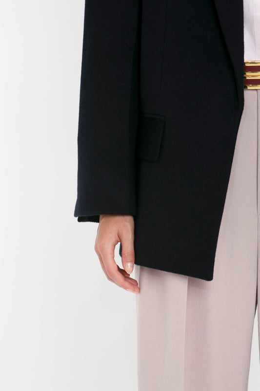 Close-up of a person wearing a Victoria Beckham Peak Lapel Jacket In Midnight, a white shirt with a gold belt, and light pink trousers. Only the lower part of the blazer, the belt, and the trousers, along with a hand, are visible, showcasing a contemporary feel reminiscent of Victoria Beckham's style.