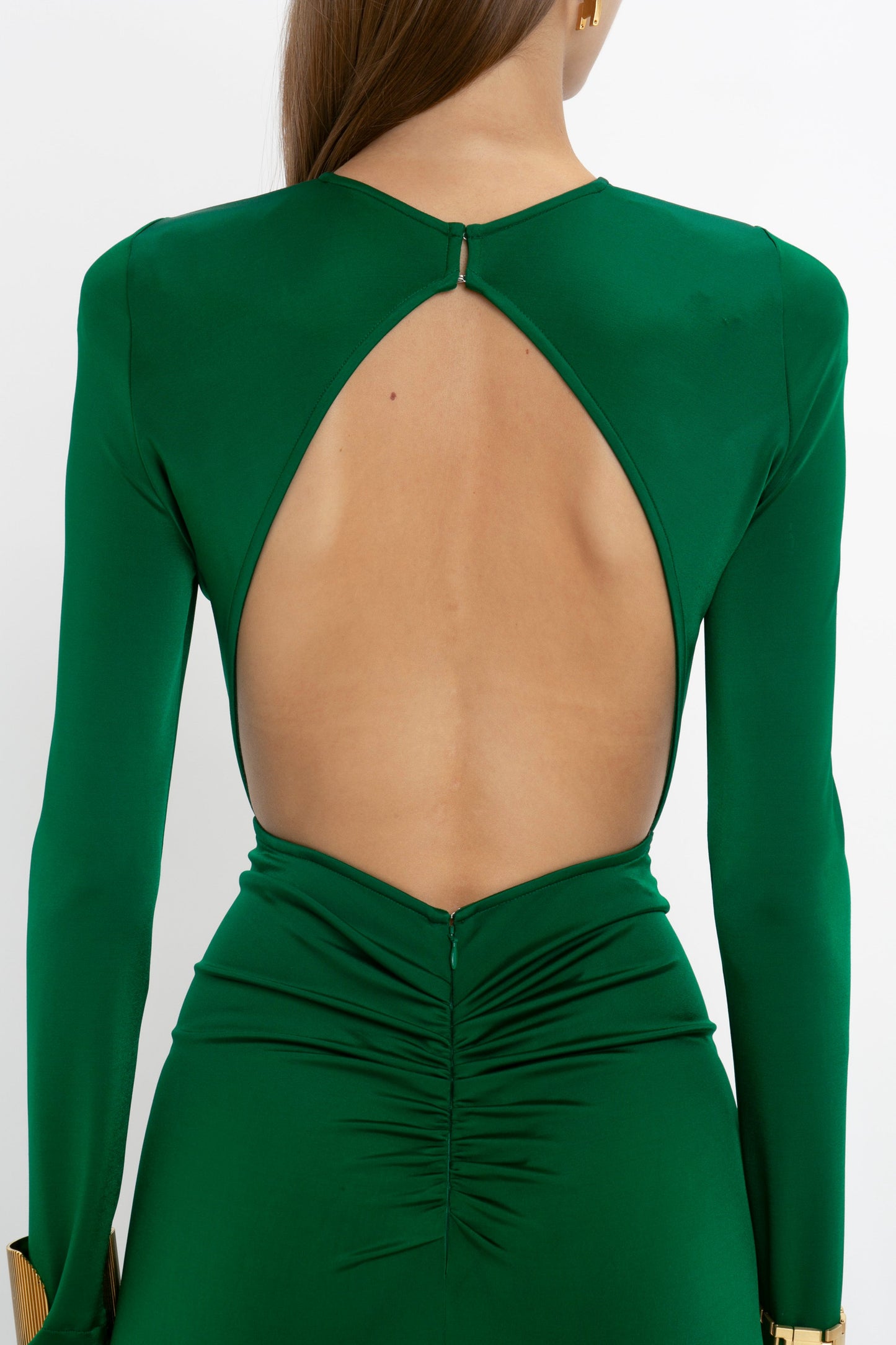 A person wearing the Victoria Beckham Circle Detail Open Back Gown In Emerald with long sleeves, ruched detailing at the lower back, and a longer-length hem is shown from behind.