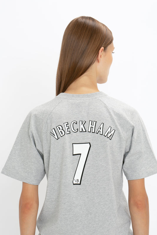 Person with long, straight hair is wearing a Victoria Beckham Football T-Shirt In Grey Marl made from organic cotton, featuring the "VBECKHAM" logo and the number "7" printed on the back. They are facing away from the camera.
