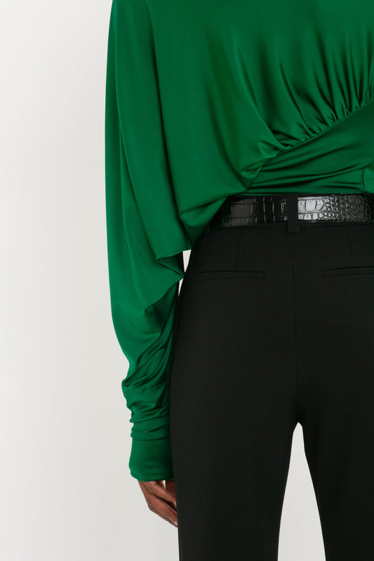 A person stands with their back to the camera, wearing a green long-sleeved blouse and contemporary Victoria Beckham Cropped Kick Trouser In Black with a black belt.