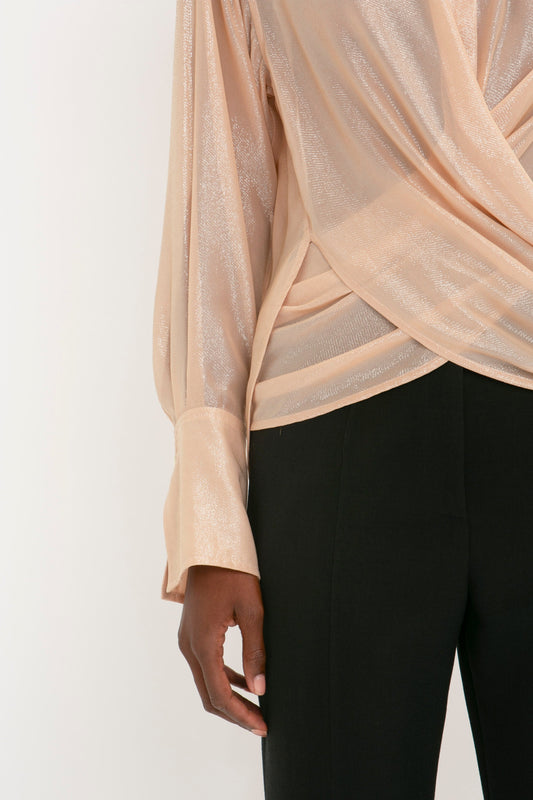 A person is shown from the shoulder to the thigh wearing a shimmery, long-sleeved Victoria Beckham Wrap Front Blouse In Rosewater with an oversized fit and black pants on a plain white background.