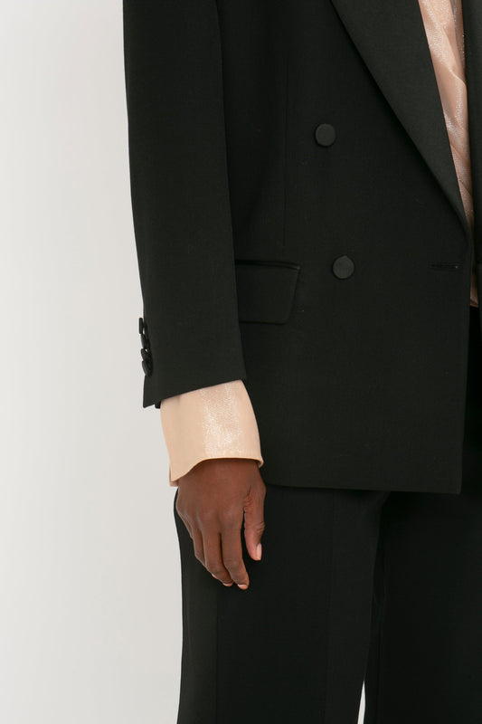 Close-up of a person in an oversized black Victoria Beckham Satin Lapel Tuxedo Jacket and pants, with a beige shirt cuff slightly exposed at the wrist. The person is standing against a plain white background.