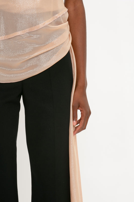 Person in black Satin Panel Straight Leg Trousers and a glittery, sheer beige Victoria Beckham Flower Detail Cami Top In Rosewater stands against a white background, with a piece of fabric hanging down on one side.