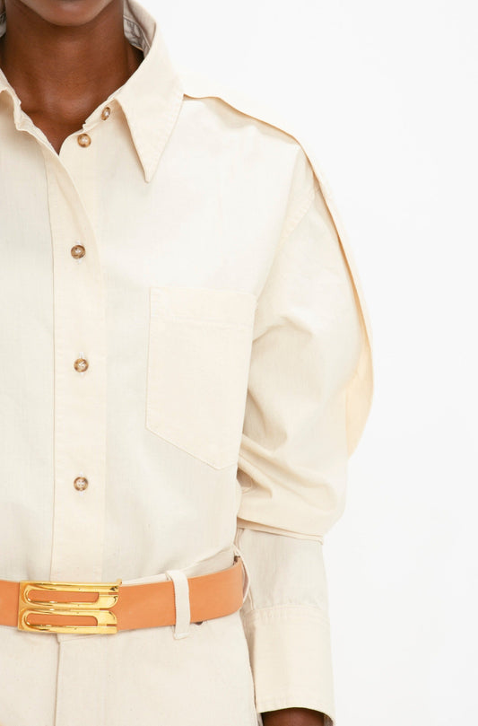 Close-up of a person wearing an Oversized Pleat Detail Denim Shirt in Ecru by Victoria Beckham with a pointed collar, a chest pocket, and a tan belt with a gold buckle. The shirt features an oversized fit and has a single shoulder epaulet.