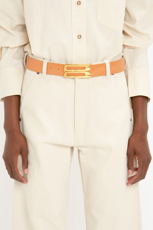Person wearing a beige button-up shirt and matching Victoria Beckham Relaxed Fit Low-Rise Jean in Ecru with a beige belt featuring a double gold buckle, showcasing a modern silhouette crafted from breathable cotton.