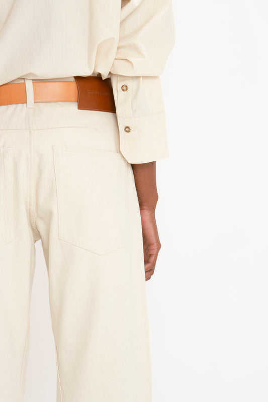 Close-up of a person wearing cream-colored pants and a matching shirt, with a light brown belt. The modern silhouette highlights the Victoria Beckham Relaxed Fit Low-Rise Jean in Ecru. Only the back and arm of the person are visible against a plain white background.
