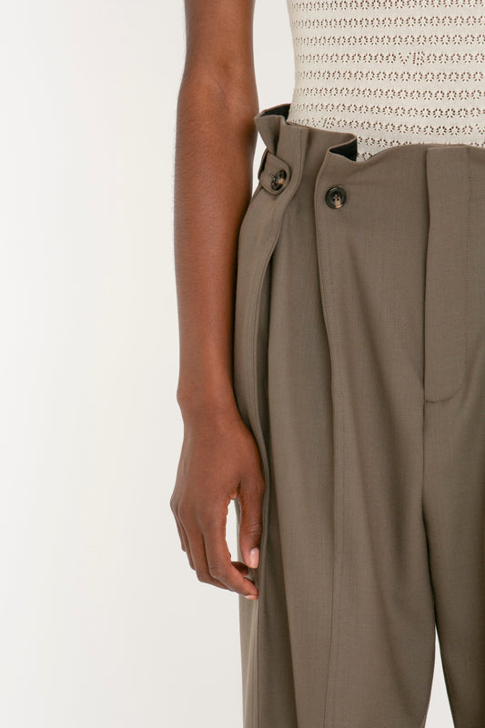 Close-up of a person wearing Victoria Beckham Gathered Waist Utility Trouser In Oregano, paired with a knitted light-colored top. The person’s left arm is relaxed by their side.