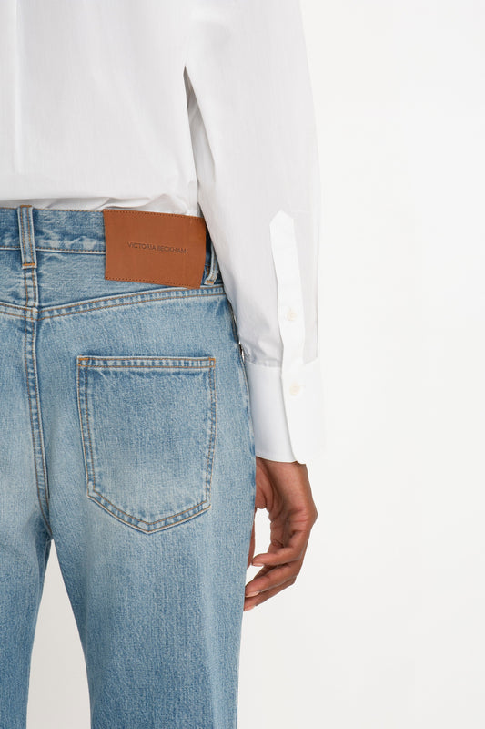 A person wearing a white long-sleeved shirt tucked into Victoria Beckham Victoria Mid-Rise Jean In Light Blue. The jeans have a brown leather patch on the waistband with the text "Victoria Beckham." The background is plain white.