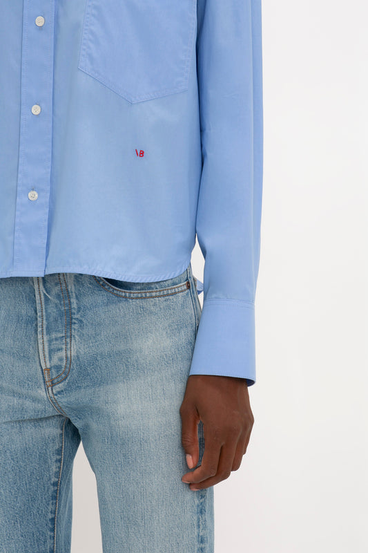 A person wearing a relaxed fit, light blue Cropped Long Sleeve Shirt In Oxford Blue by Victoria Beckham and light blue jeans. Only the lower part of the organic cotton shirt and the upper part of the jeans are visible. The person's right arm hangs by their side.