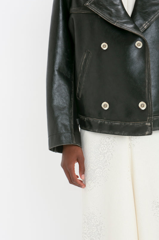 Person wearing an Oversized Leather Jacket In Black by Victoria Beckham with a distressed finish over a white lace outfit. Only the lower half of the jacket and the person's hand are visible.