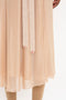 Close-up of a person wearing a sheer, beige dress with a wrap front design and two beige ties hanging down, featuring small red letters "V" and "B" at the ends. The Victoria Beckham Wrap Front Cardigan In Nougat is partially transparent, revealing the legs underneath.