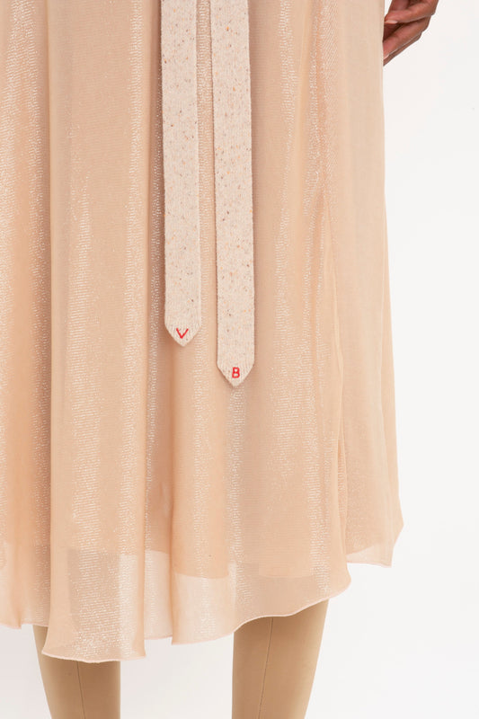 Close-up of a person wearing the Victoria Beckham Flower Detail Cami Skirt in Rosewater with a shimmery texture and long beige ties hanging down. Only the lower part of the fit-and-flare silhouette and legs in beige leggings are visible.