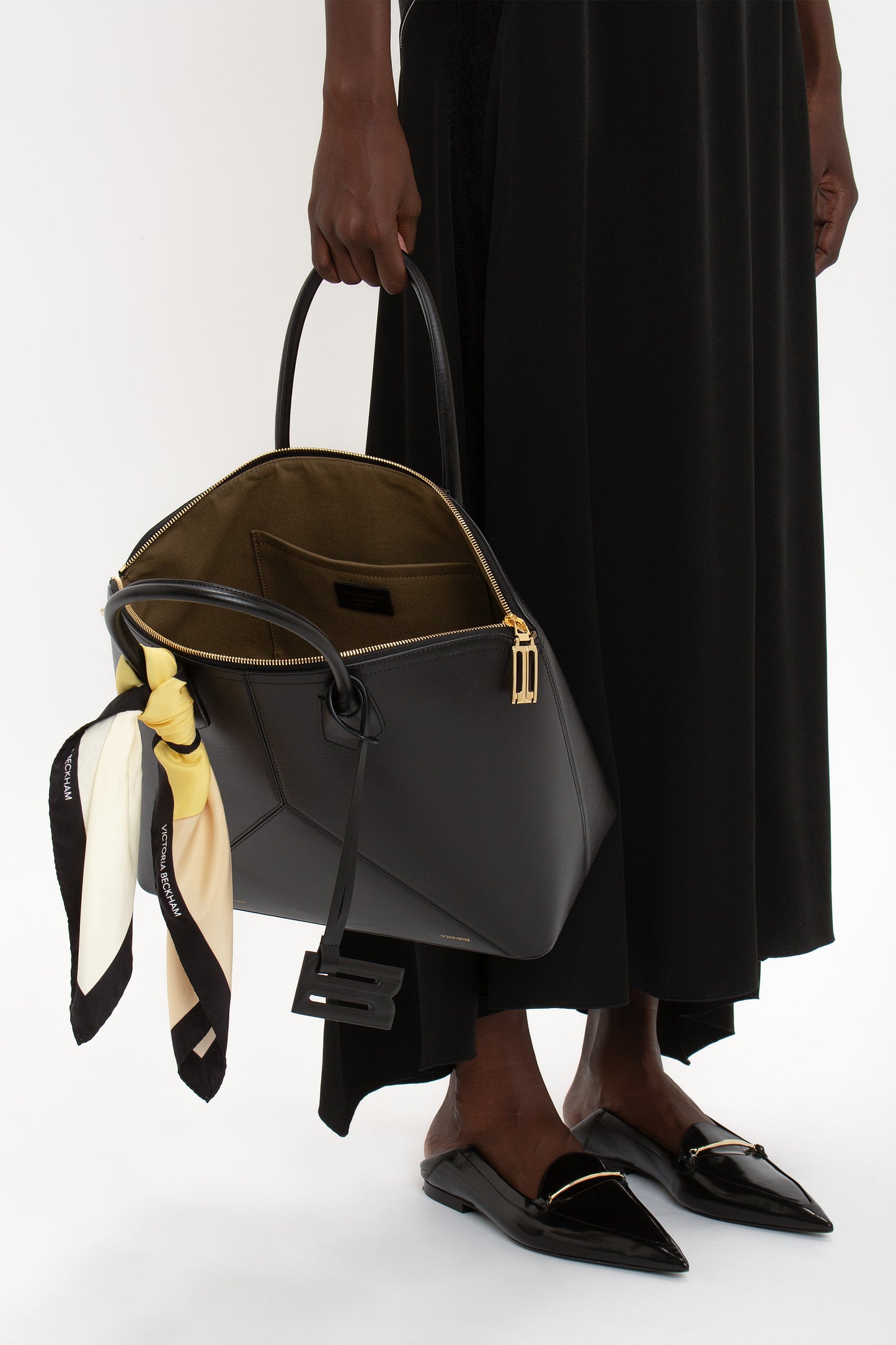 Person holding a Victoria Beckham Victoria Bag In Black Leather tied with a yellow and white scarf, wearing a black dress and black pointed shoes.