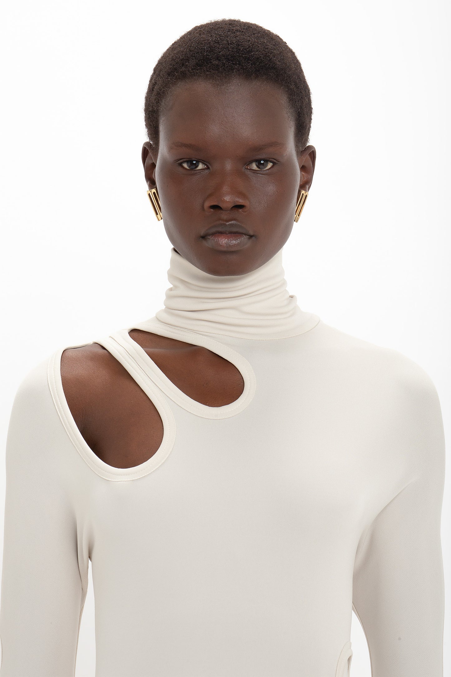 A person with short hair is wearing a high-neck cream-colored top with unusual cutouts near the shoulder and gold hoop earrings, staring directly at the camera. The background is plain white, giving focus to the liquid crepe jersey fabric of their *Long Sleeve Cut-Out Jersey Midi Dress In Bone* from *Victoria Beckham*.