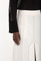 A close-up of a person wearing the Victoria Beckham Pocket Detail Shirt In Black and a white skirt with vertical slits and intricate stitching details. The person's hand is visible resting by their side, emphasizing the tailored design details of the ensemble.