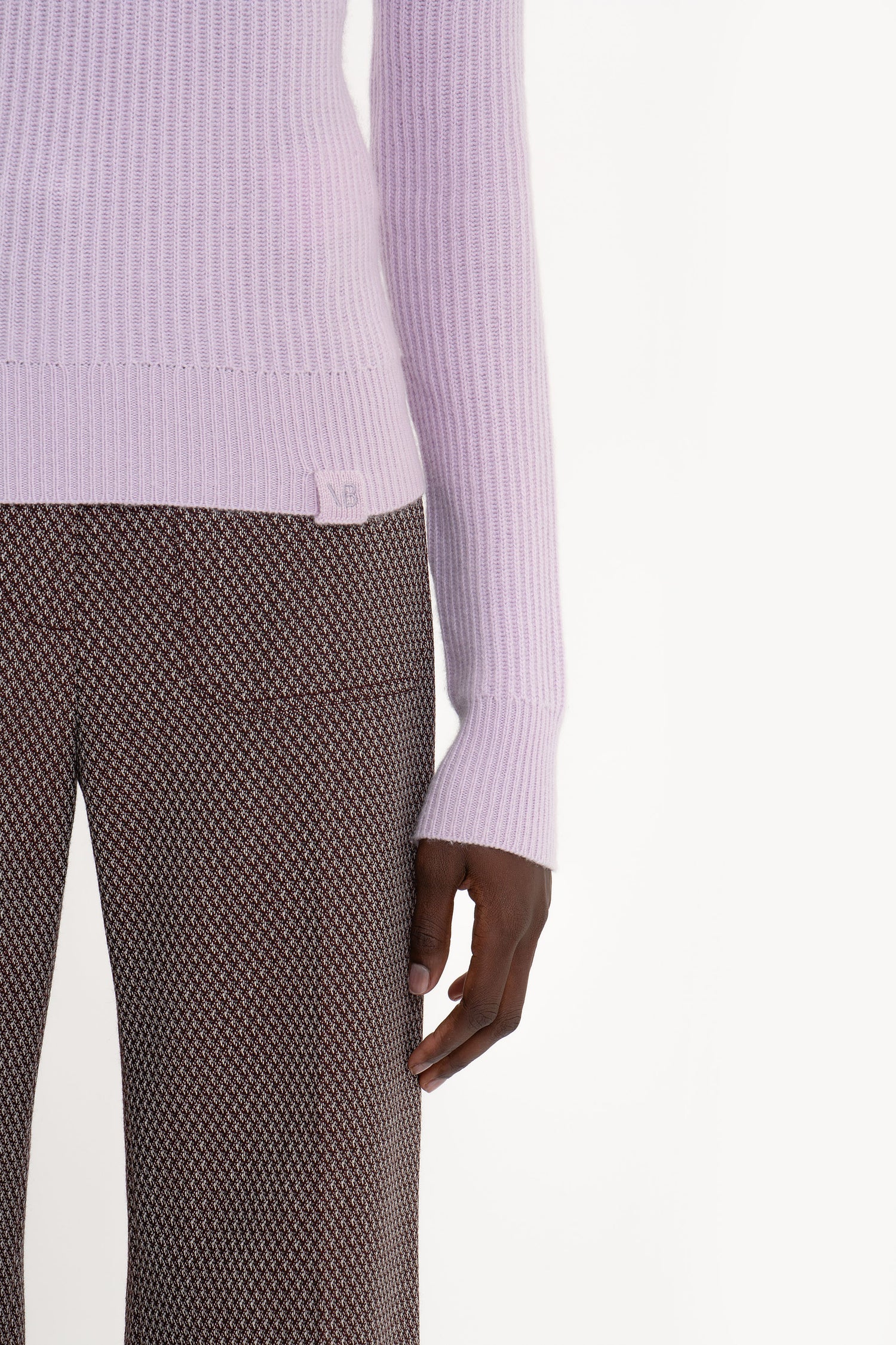 A person wearing a light purple, fisherman’s ribbed knit Victoria Beckham Double Collared Jumper In Petunia and brown patterned pants, with their left arm relaxed at their side.