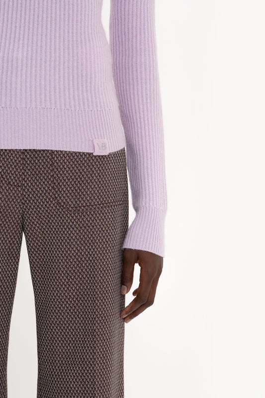 A person wearing a light purple, fisherman’s ribbed knit Victoria Beckham Double Collared Jumper In Petunia and brown patterned pants, with their left arm relaxed at their side.