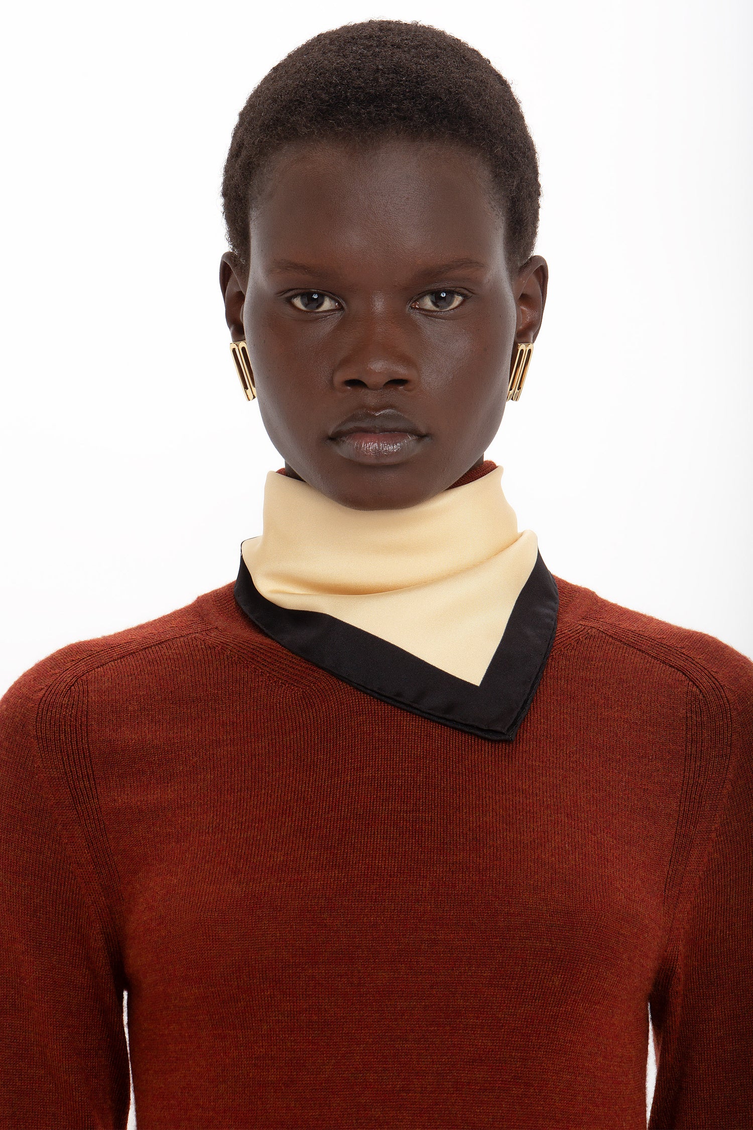 A person with short hair wearing a rust-colored sweater, a beige and black scarf, gold earrings, and a Victoria Beckham High Neck Tie Detail Dress In Russet.