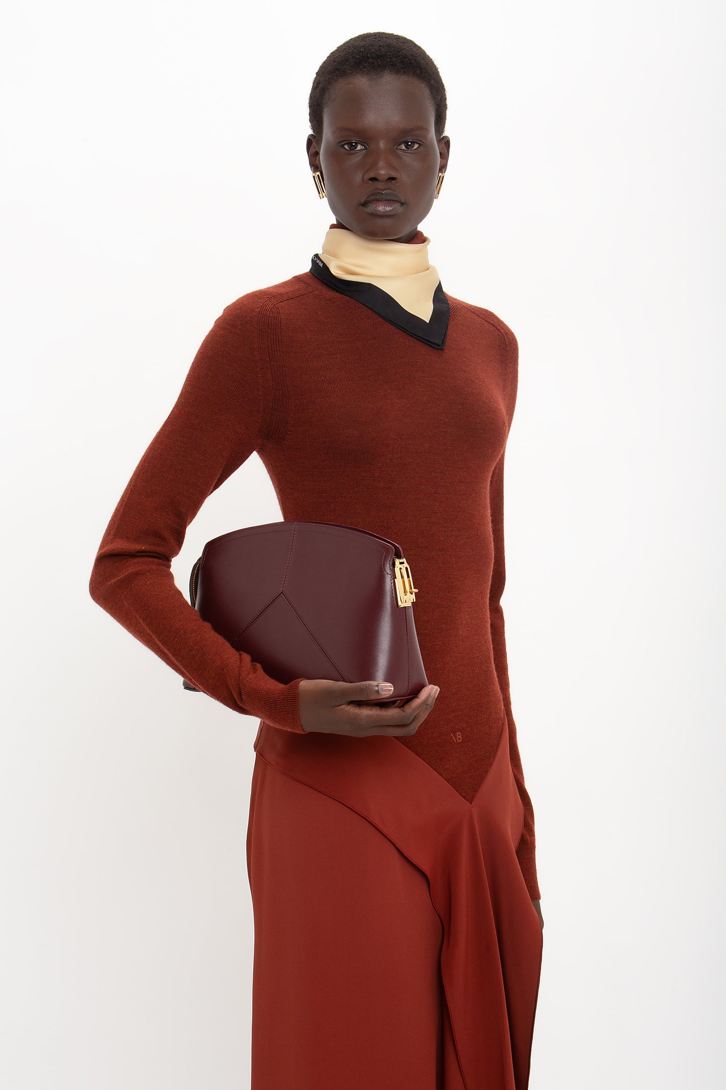 A person poses in a High Neck Tie Detail Dress In Russet, reminiscent of a Victoria Beckham collection, holding a maroon handbag against a plain white background.
