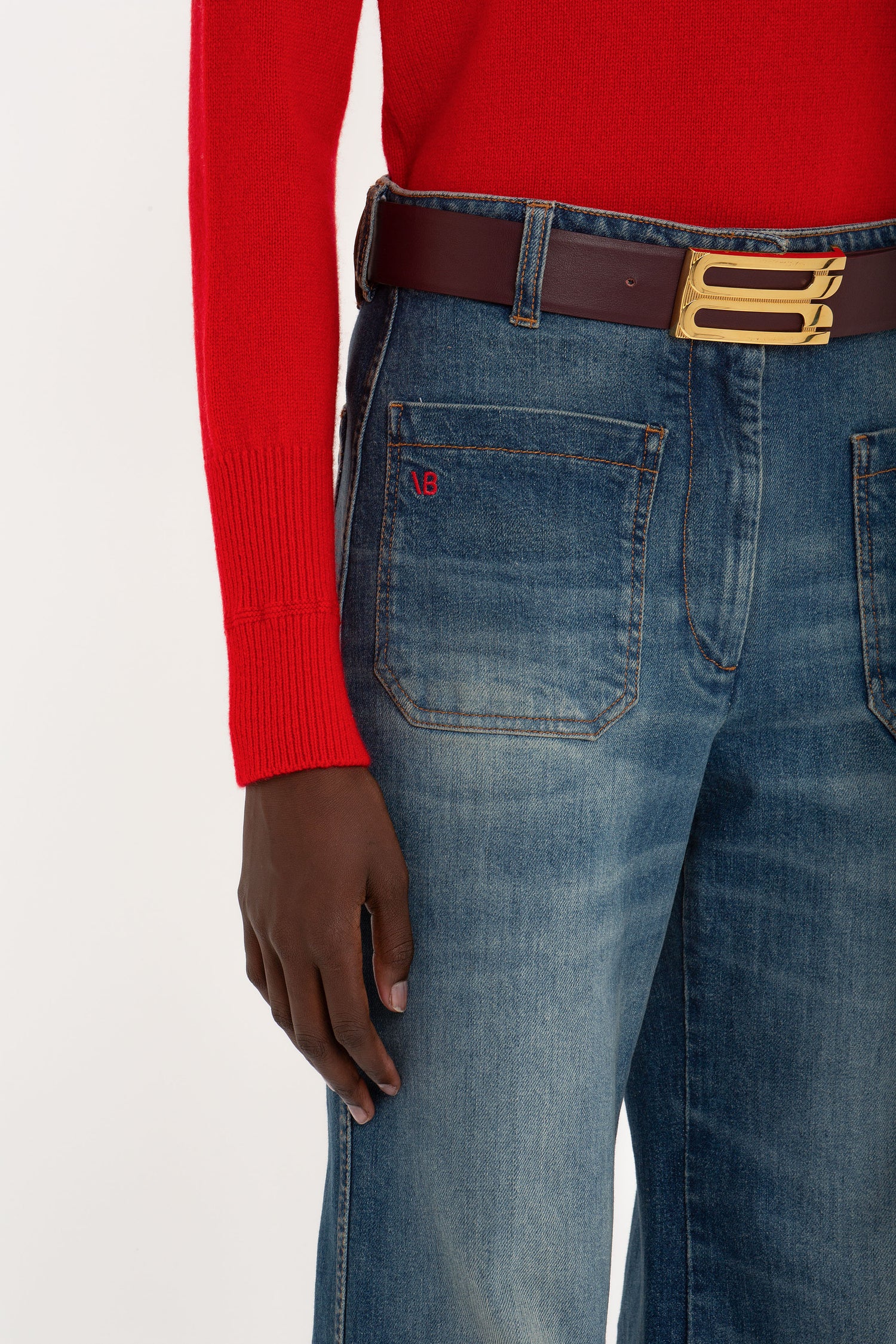 A person wearing a red long-sleeve shirt, blue denim jeans with a brown belt, and a gold buckle. The Victoria Beckham Alina High Waisted Jean In Heavy Vintage Indigo Wash are complemented by the person's right hand placed near the right pocket.