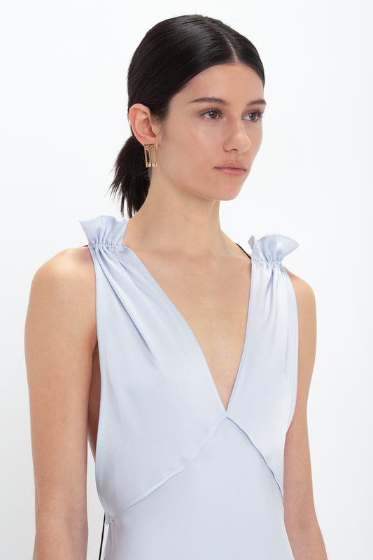 A person with dark hair tied back is wearing the Victoria Beckham Exclusive Gathered Shoulder Cami Floor-Length Gown In Ice Blue. The light blue sleeveless design features tied shoulder straps and a deep V-neckline, reminiscent of a gathered shoulder cami gown.