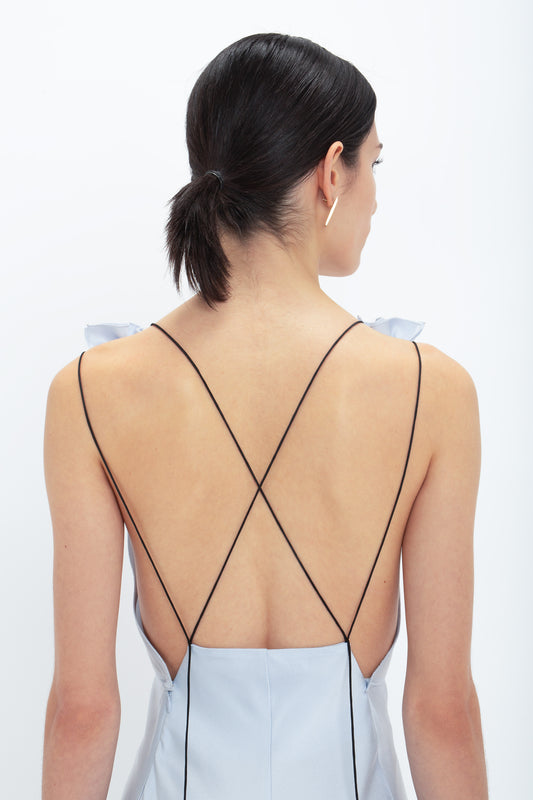 A person with dark hair in a low ponytail wears an ice blue, backless dress with thin black straps forming an X on the back. The Victoria Beckham Exclusive Gathered Shoulder Cami Floor-Length Gown In Ice Blue, made from crepe back satin, drapes elegantly as they face away from the camera.