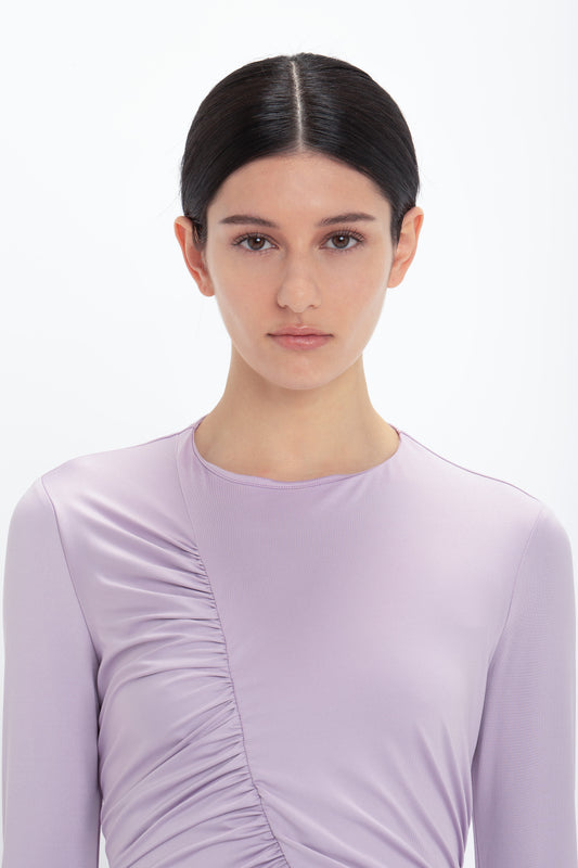 A woman with dark hair pulled back, wearing a Victoria Beckham Ruched Detail Floor-Length Gown In Petunia, exudes understated glamour as she stands against a white background, looking directly at the camera.