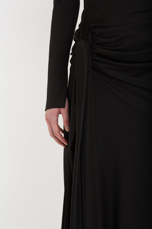 Sentence with replacement: A close-up of a person wearing a Victoria Beckham black gown with a detailed ruched design on the side. Only the lower torso and hand are visible.