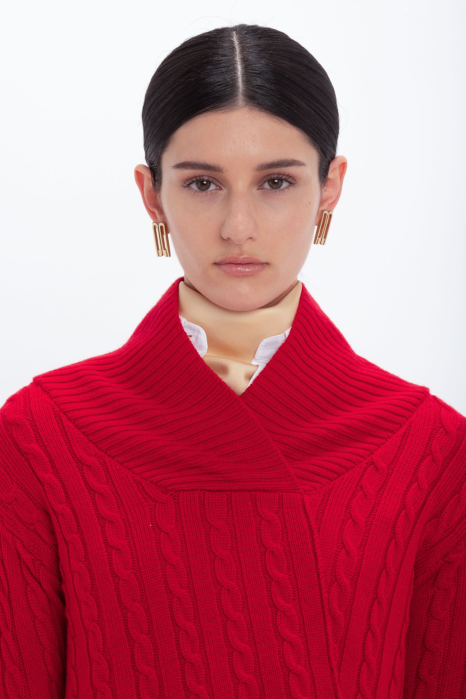 A person with straight dark hair pulled back, wearing gold earrings and a luxurious red, Victoria Beckham Wrap Detail Jumper In Red with a high collar, stands against a plain white background.