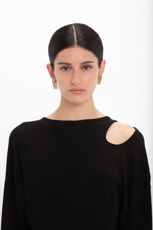 A woman with dark hair styled in a center part, wearing a black Victoria Beckham Twist Detail Jersey Top with shoulder cut-outs and gold earrings, facing the camera with a neutral expression.