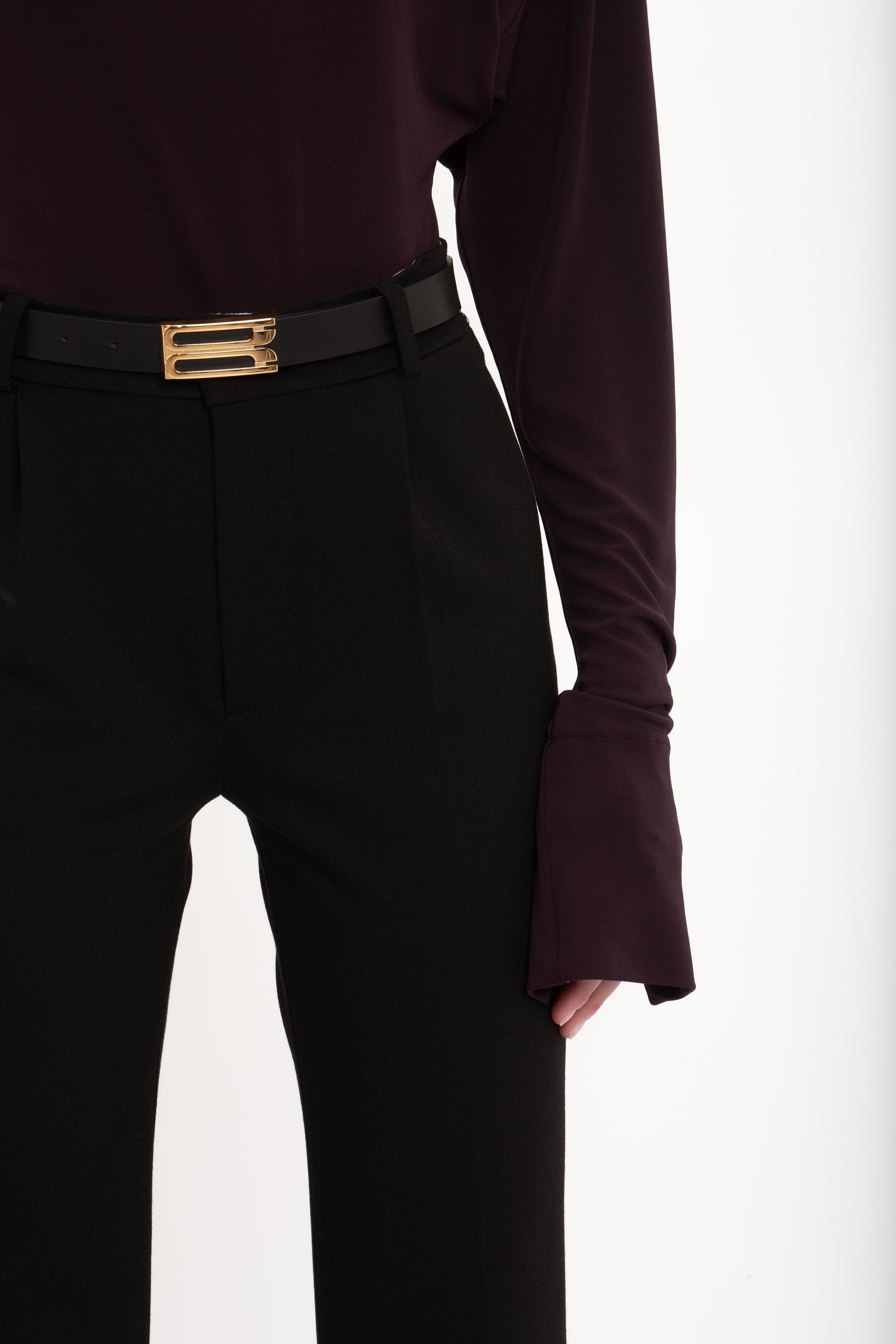 A person wearing a dark long-sleeve top and Cropped Kick Trouser In Black by Victoria Beckham with a black belt featuring a gold buckle, embracing a minimalist aesthetic.