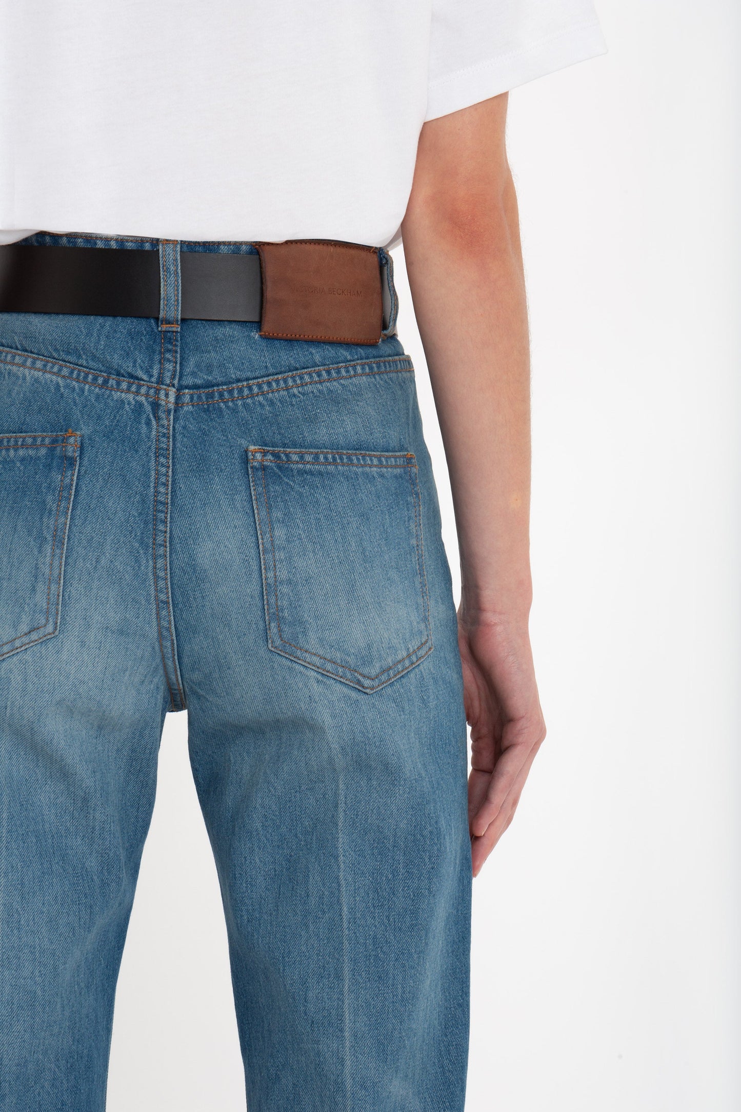 Back view of a person wearing a white t-shirt and blue jeans with a brown belt; their right arm is relaxed by their side. The high waistline of the 100% cotton Julia Jean In Broken Vintage Wash by Victoria Beckham adds a stylish touch to the outfit.