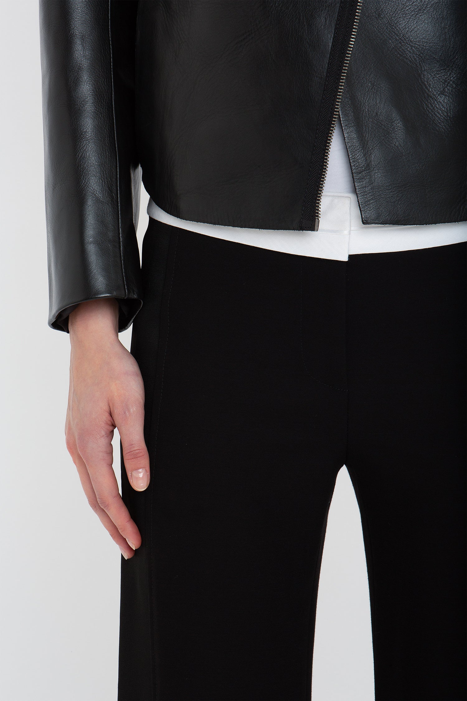 Close-up of a person wearing a Victoria Beckham Tailored Leather Biker Jacket In Black, a white shirt, and black pants, with their left hand resting at their side.