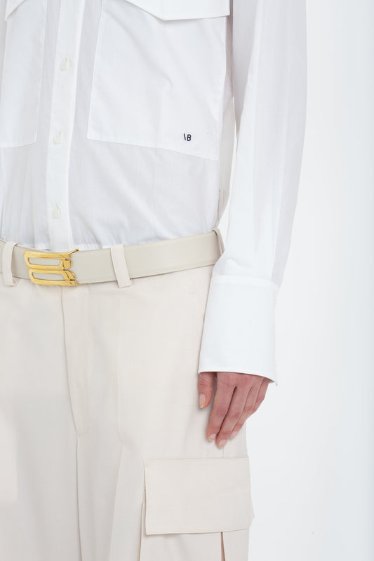 Close-up of a person wearing a white button-up shirt, beige belt with a yellow buckle, and Victoria Beckham's Relaxed Cargo Trouser In Bone with a pocket flap. The military-inspired shirt has large cuffs and a small logo.