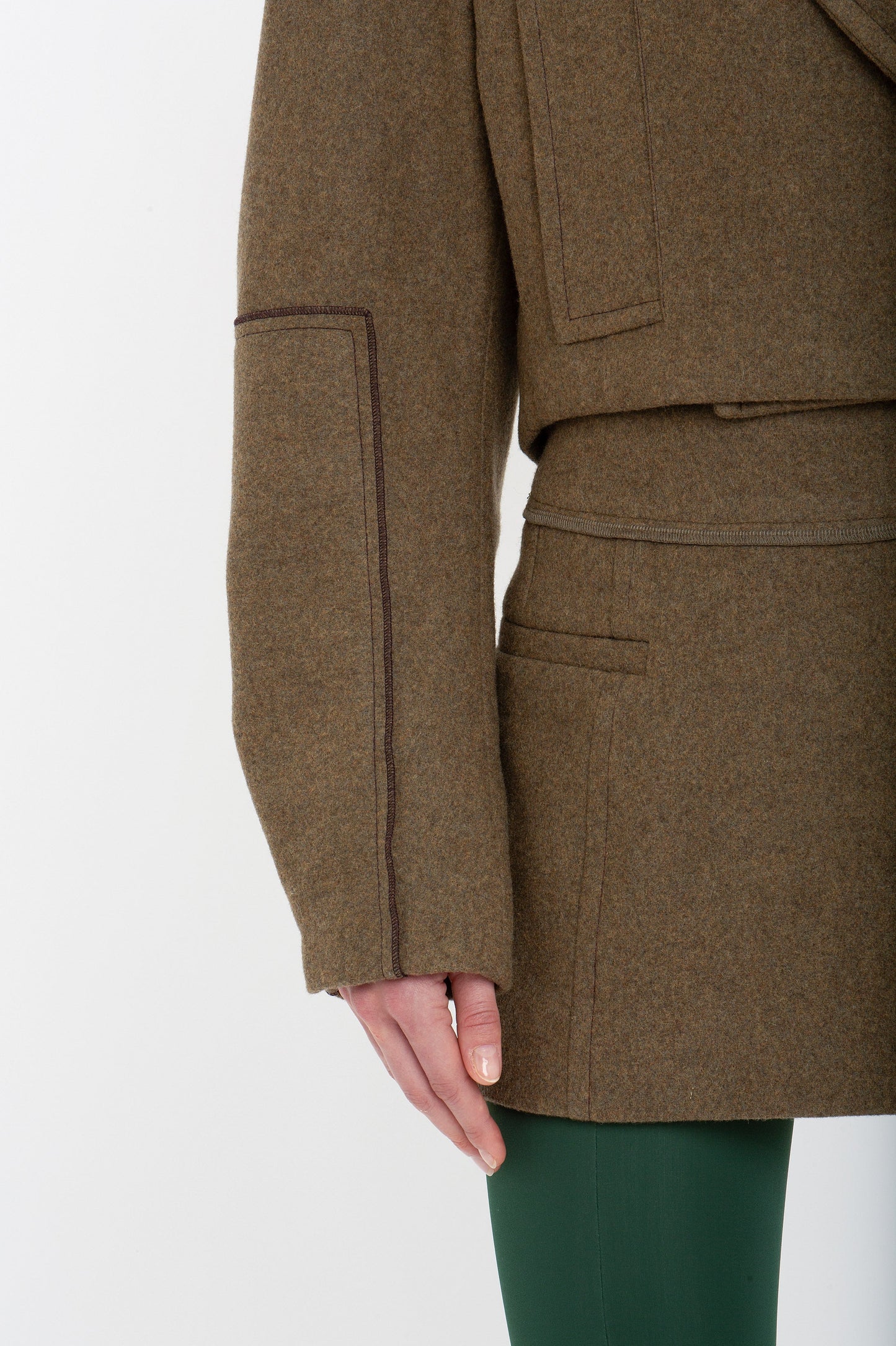 Close-up of a person wearing a Tailored Mini Skirt In Khaki by Victoria Beckham and green pants, showing the side seam and pocket detail of the coat, with their left arm resting at their side.