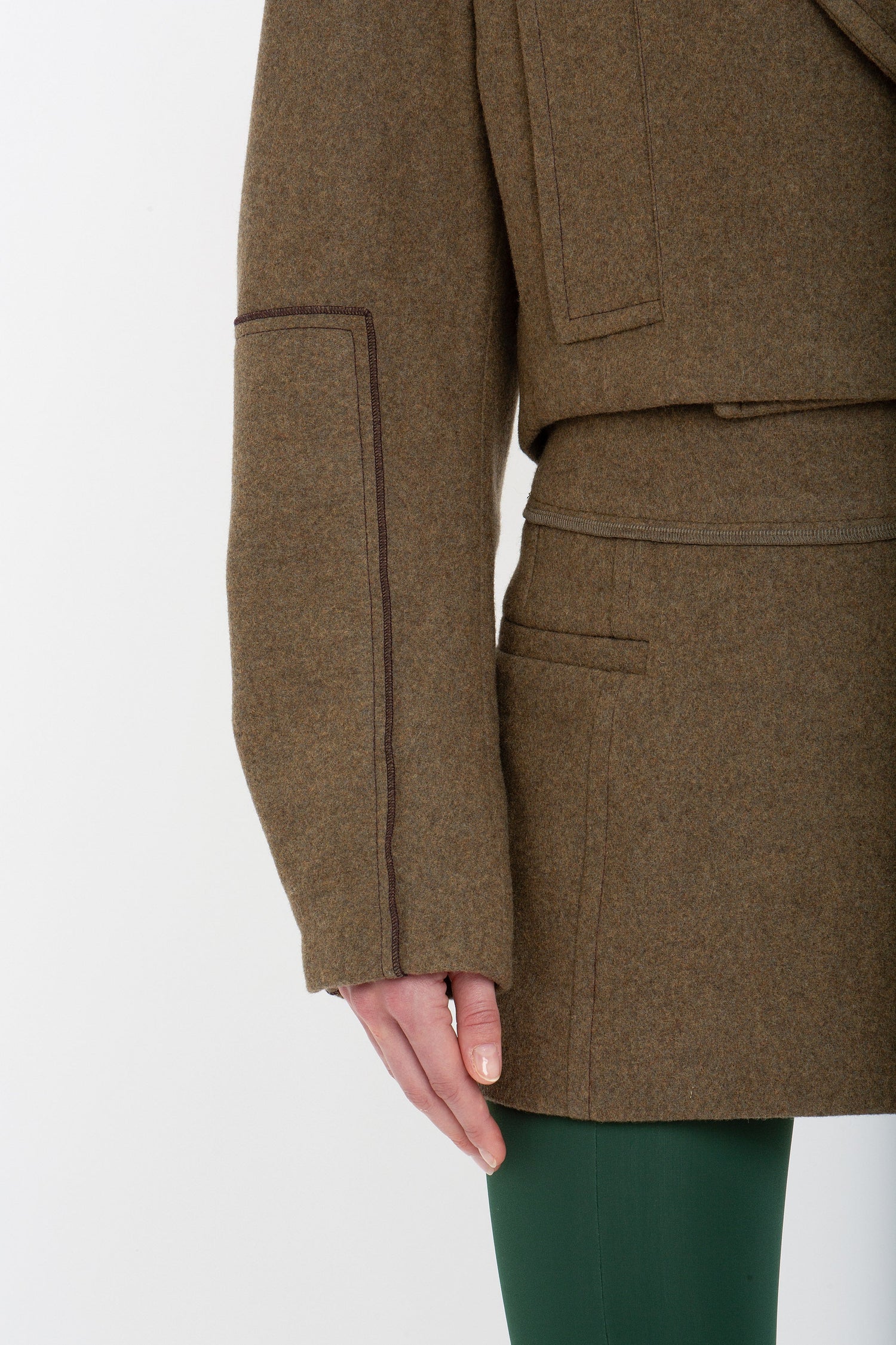 Close-up of a person wearing a Tailored Mini Skirt In Khaki by Victoria Beckham and green pants, showing the side seam and pocket detail of the coat, with their left arm resting at their side.
