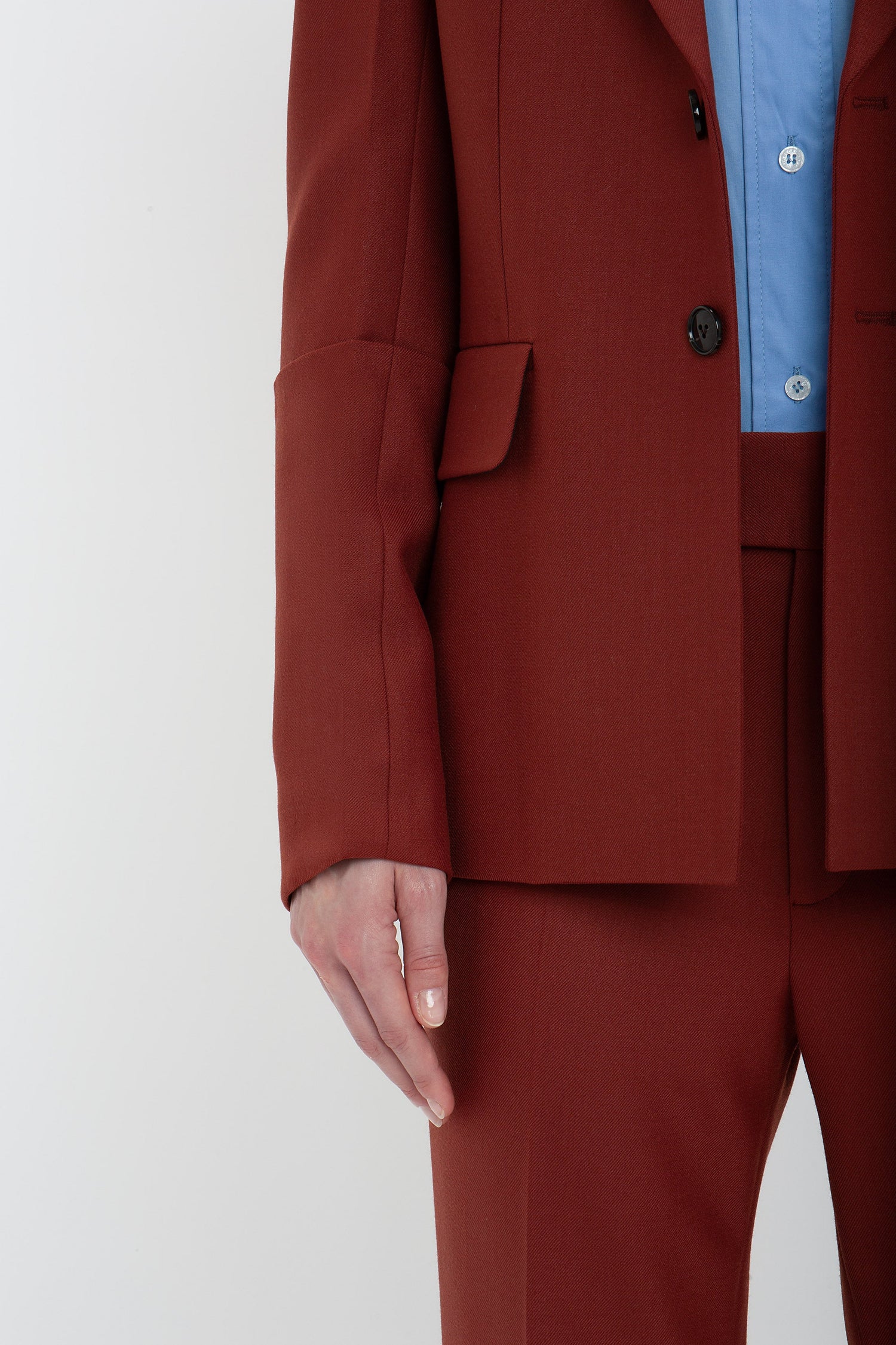 Person wearing a Victoria Beckham Sleeve Detail Patch Pocket Jacket In Russet with contemporary detailing, paired with a blue shirt. They show their left arm and a portion of their torso, focusing on the lower half of the jacket and part of the pants.