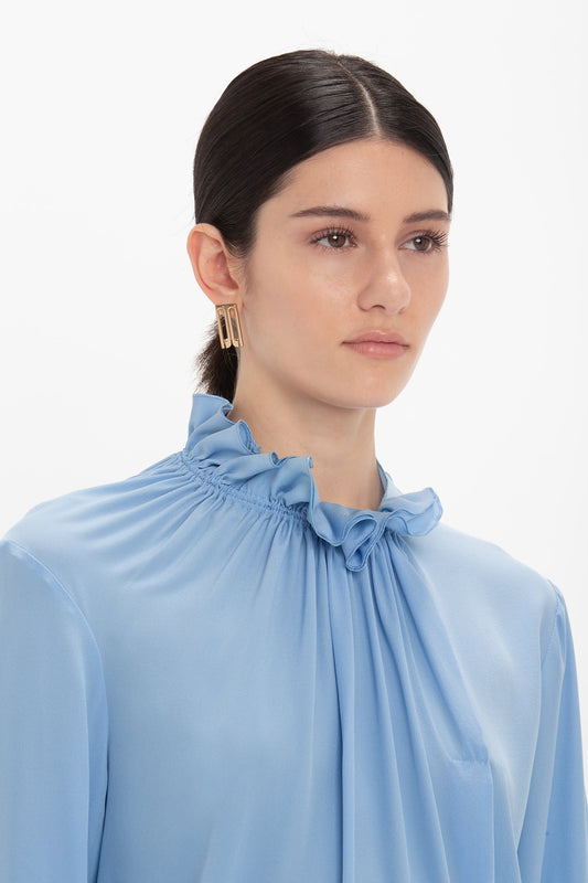 A person with long dark hair tied back wears an Exclusive Ruffle Neck Blouse In Cornflower Blue by Victoria Beckham and large rectangular earrings.
