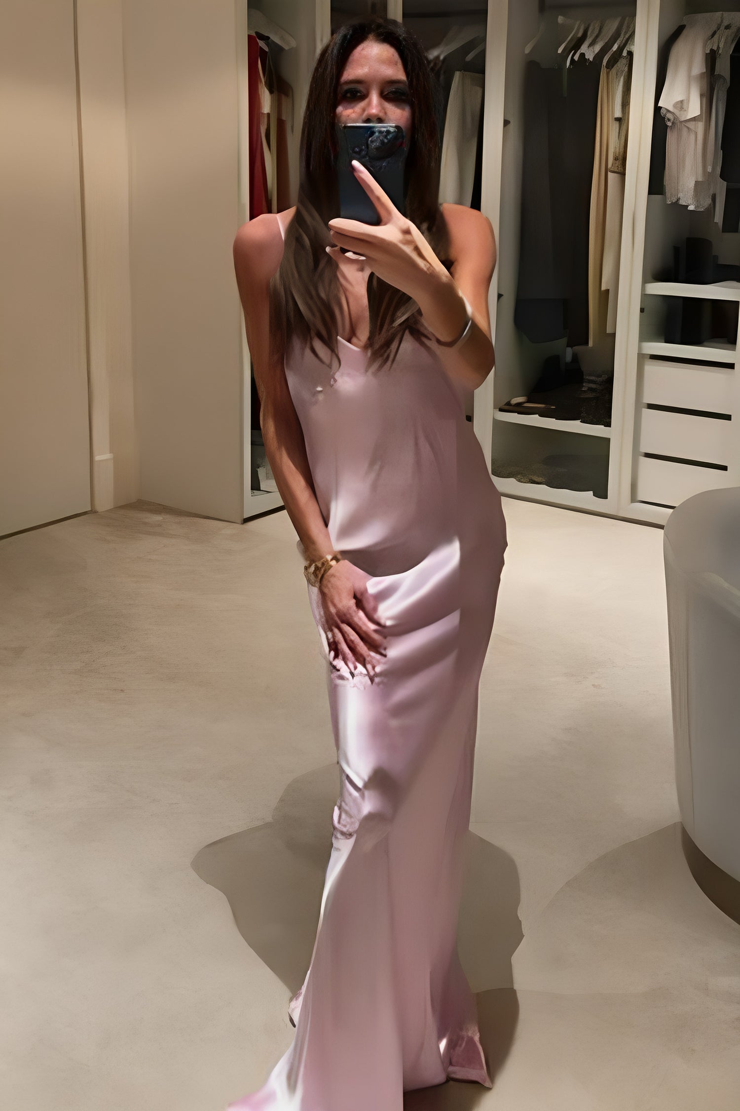 A woman takes a mirror selfie in a wardrobe, wearing a Low Back Cami Floor-Length Dress by Victoria Beckham and holding a smartphone up to her face.