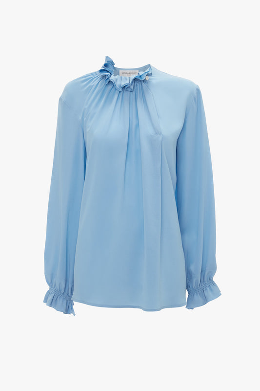 Exclusive Ruffle Neck Blouse In Cornflower Blue from Victoria Beckham, long-sleeved with a ruffled neckline and cuffs, featuring a gathered design at the front.