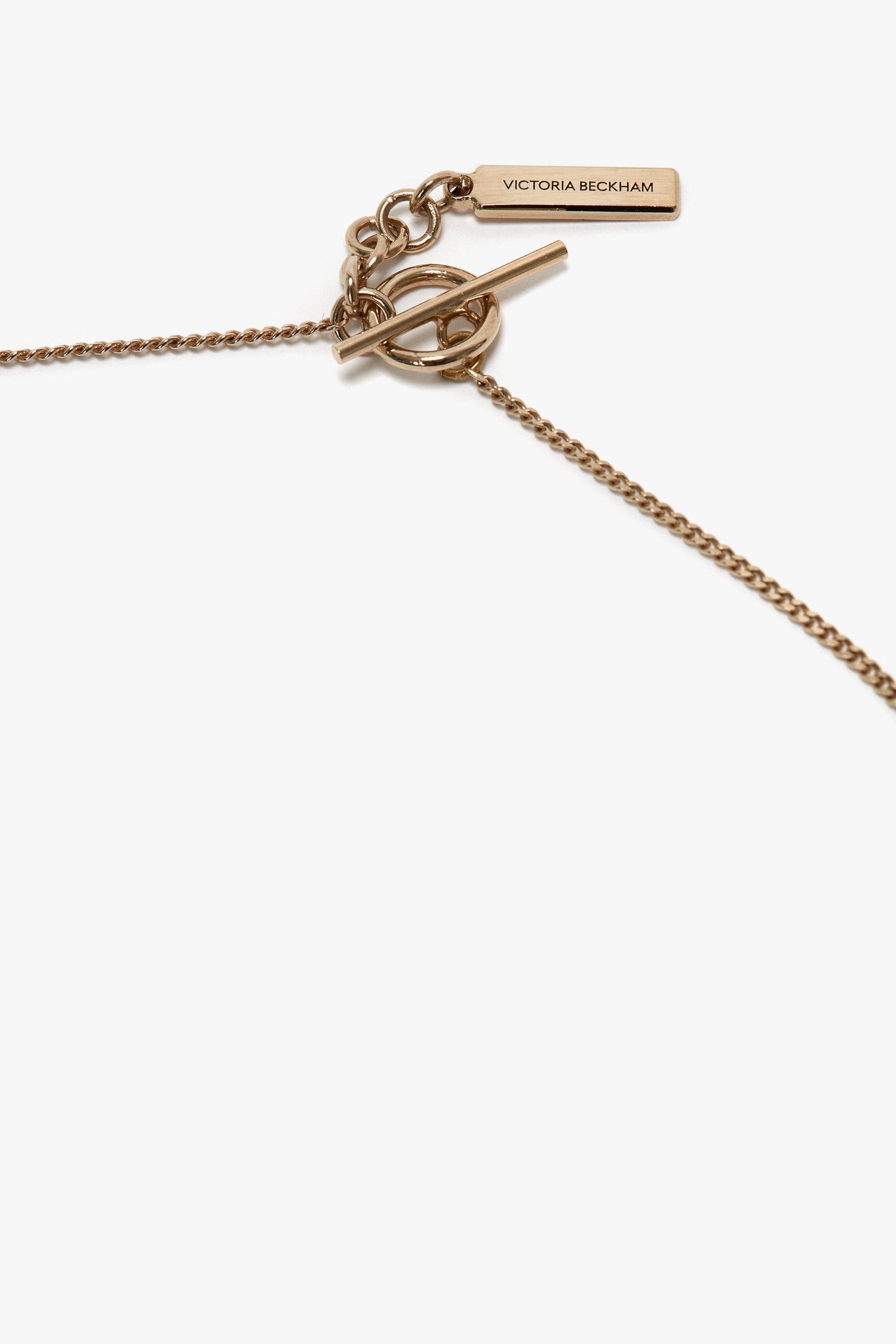 An Exclusive Body Charm Necklace In Light Gold featuring a toggle clasp and a small rectangular tag engraved with "Victoria Beckham," crafted from 100% brass.